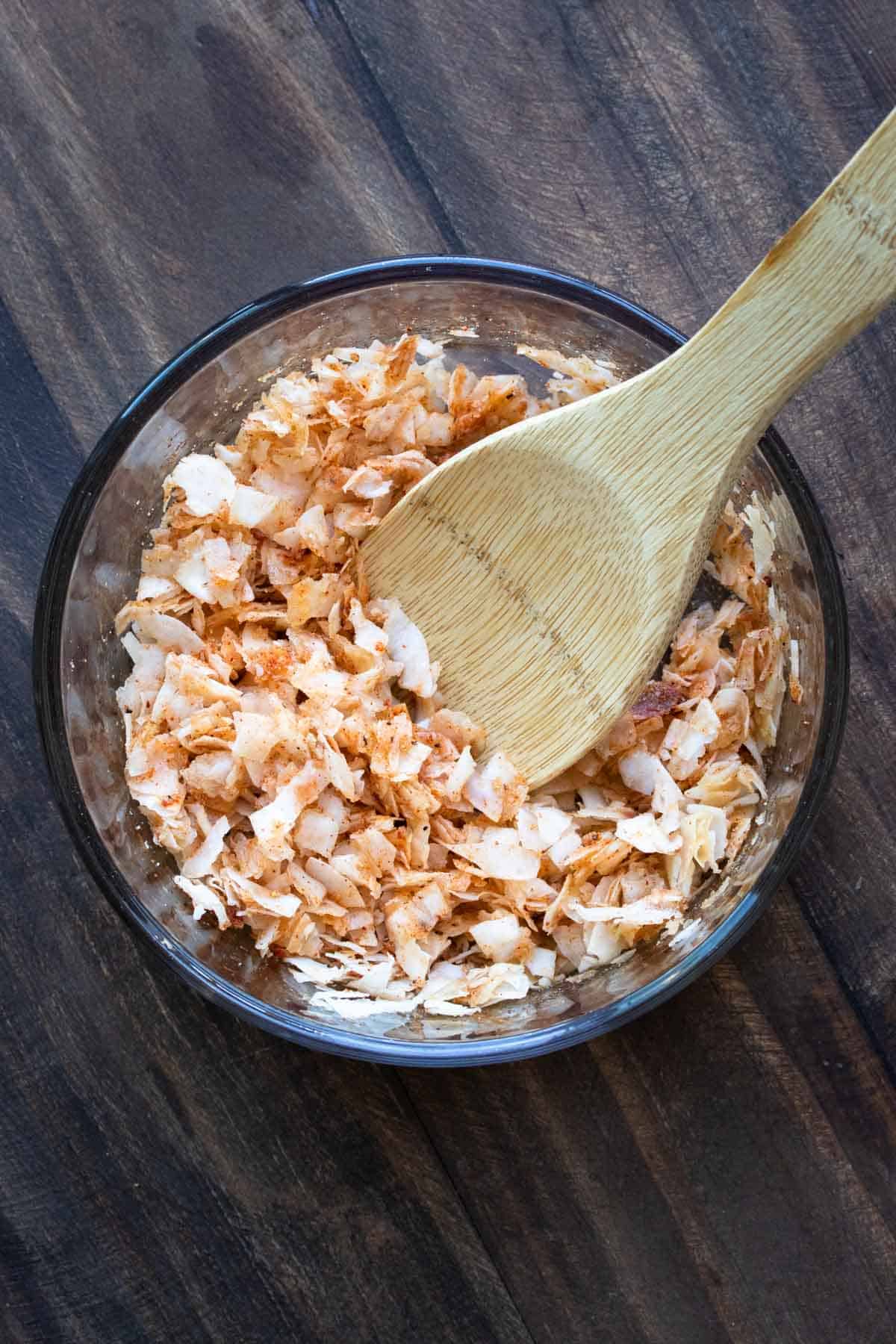 Wooden spoon mixing coconut flakes and seasonings in a glass bowl