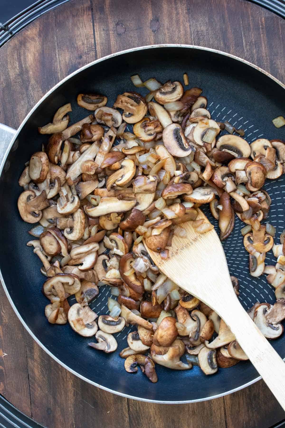 Wooden spoon mixing mushrooms and onions in a pan.