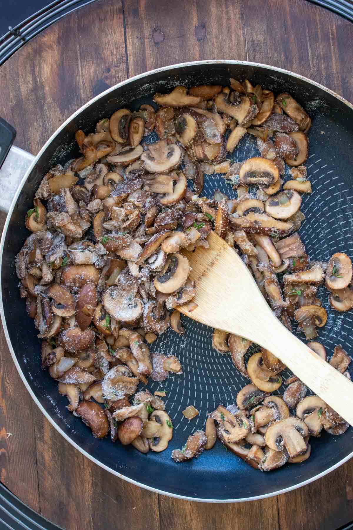 Flour coated mushrooms and onions being mixed in a pan by a wooden spoon.