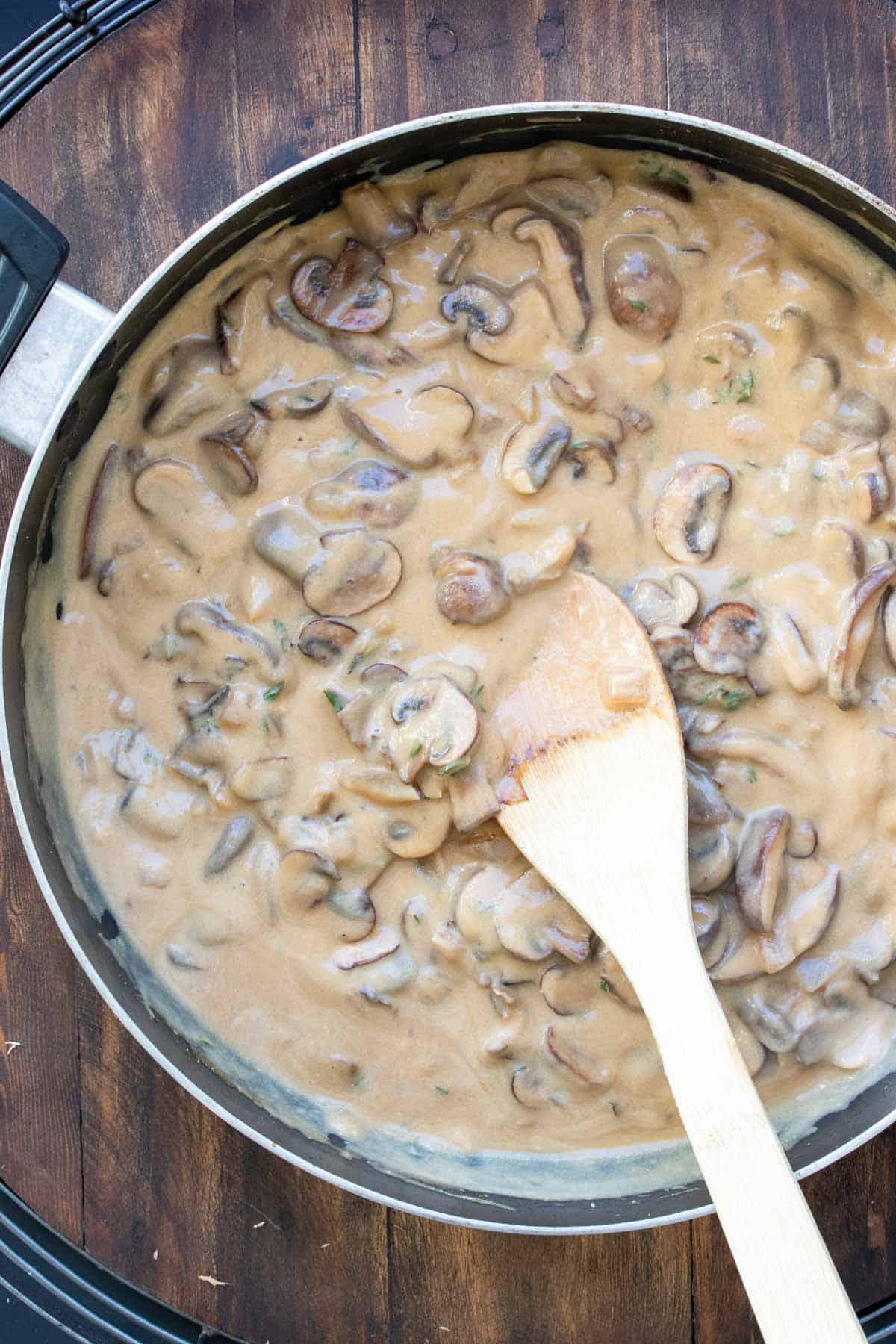 Wooden spoon mixing a tan creamy sauce with mushrooms in a pan.