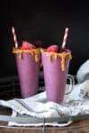 Two berry smoothies in tall glasses with a rim of peanut butter around the top and striped straws