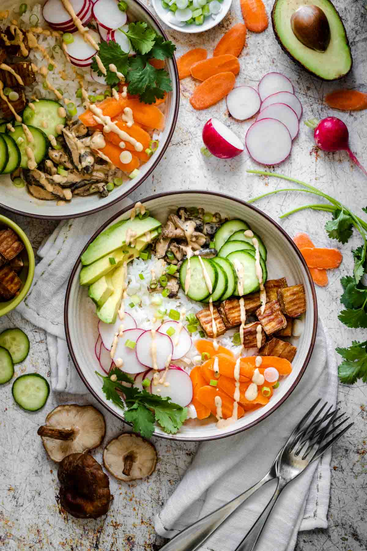 Top view of two sushi bowls surrounded by ingredients on a brown speckled white surface