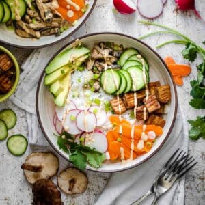 A cream bowl filled with rice topped with veggies, tofu and a pink sauce drizzled on top on a brown speckled white surface