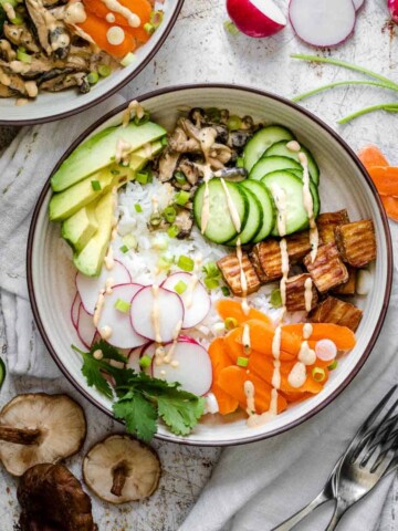 A cream bowl filled with rice topped with veggies, tofu and a pink sauce drizzled on top on a brown speckled white surface