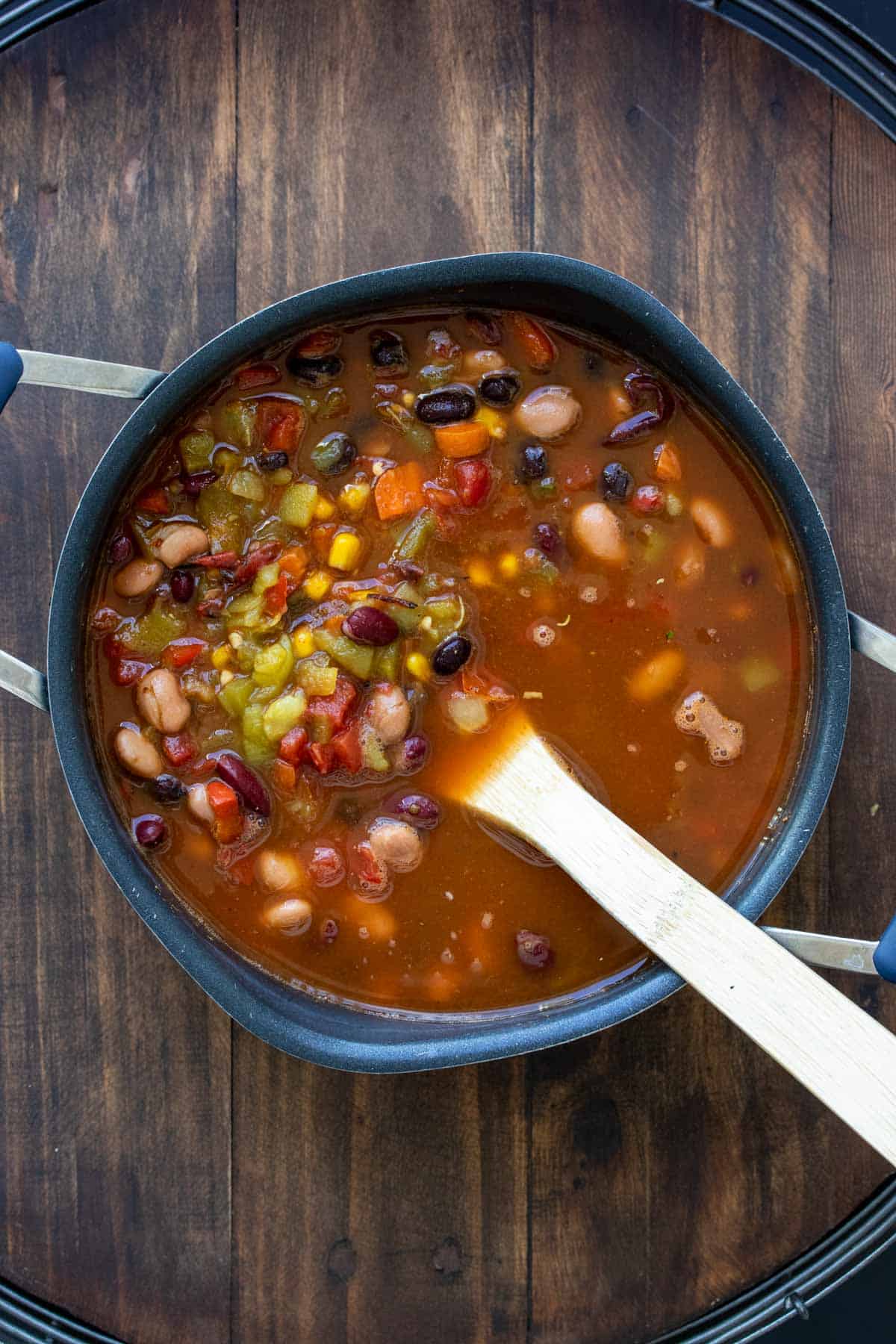 Pot filled with broth, veggies and beans that are being mixed with a wooden spoon