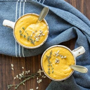 Two cream colored mugs on a wooden surface with pureed winter squash soup and spoons in them