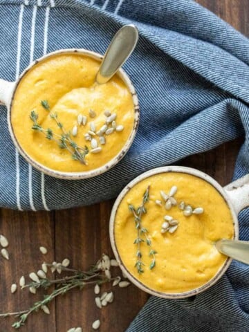 Two cream colored mugs on a wooden surface with pureed winter squash soup and spoons in them