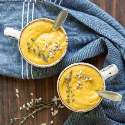 Two cream colored mugs on a wooden surface with pureed winter squash soup and spoons in them