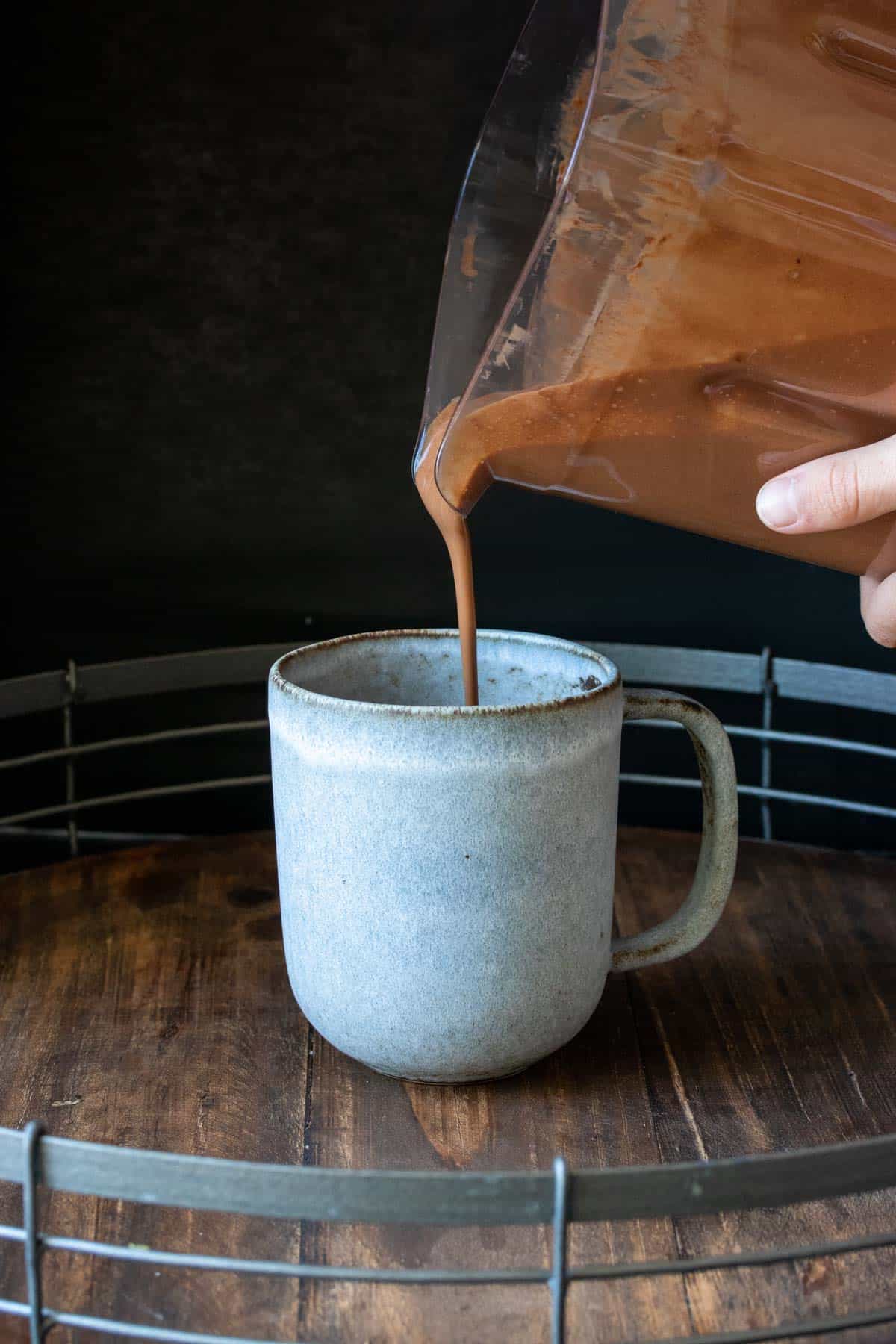 Hand pouring a thick chocolate milk into a grey mug