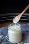 A small wooden spoon dripping coconut butter into a glass jar sitting on a blue denim towel.