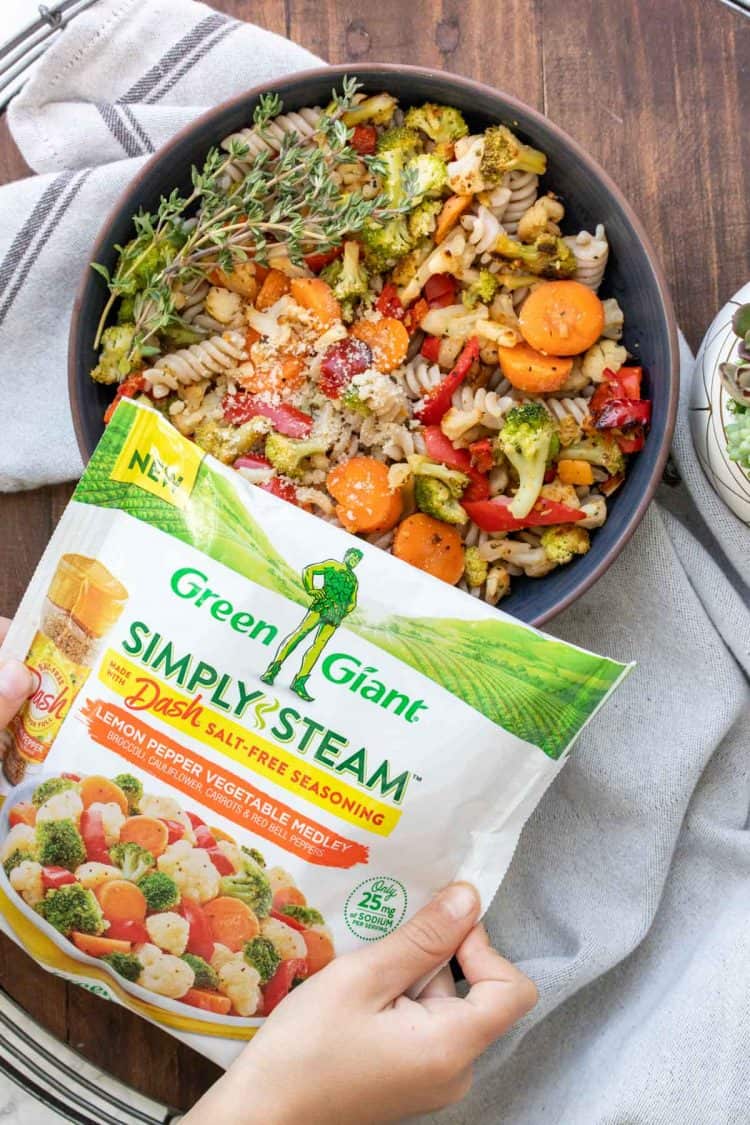 Hands holding a bag of frozen veggies over a bowl of pasta and veggies