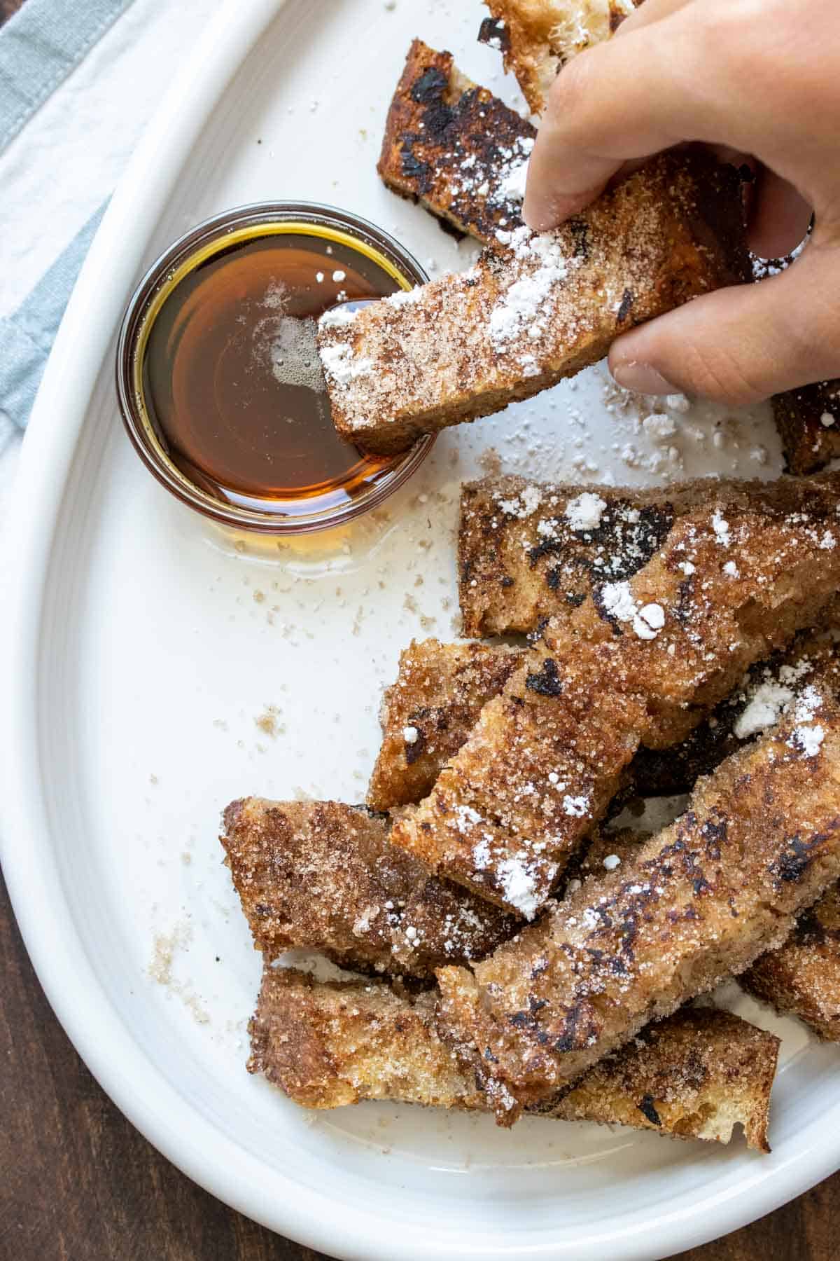 Hand dipping a french toast stick in a small bowl of maple syrup
