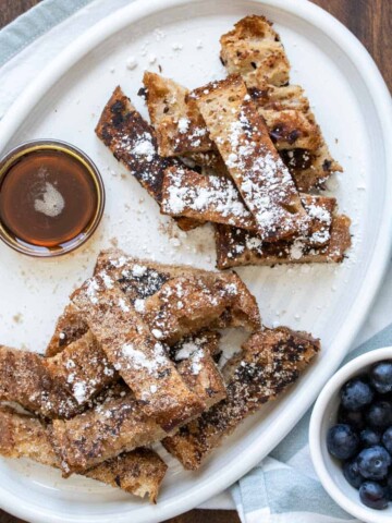 Two piles of french toast sticks on a white plater surrounded by toppings in bowls