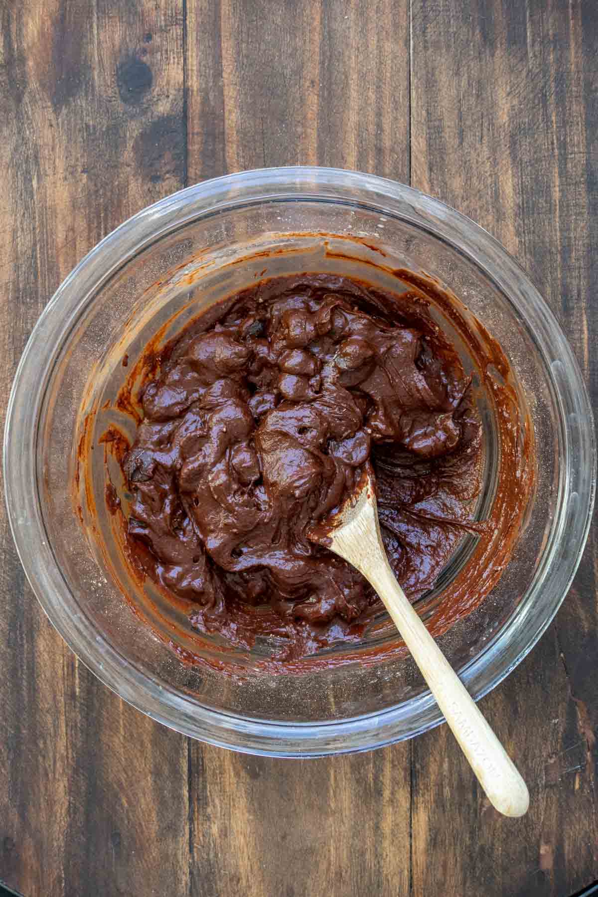 Wooden spoon mixing chocolate batter in a glass bowl.