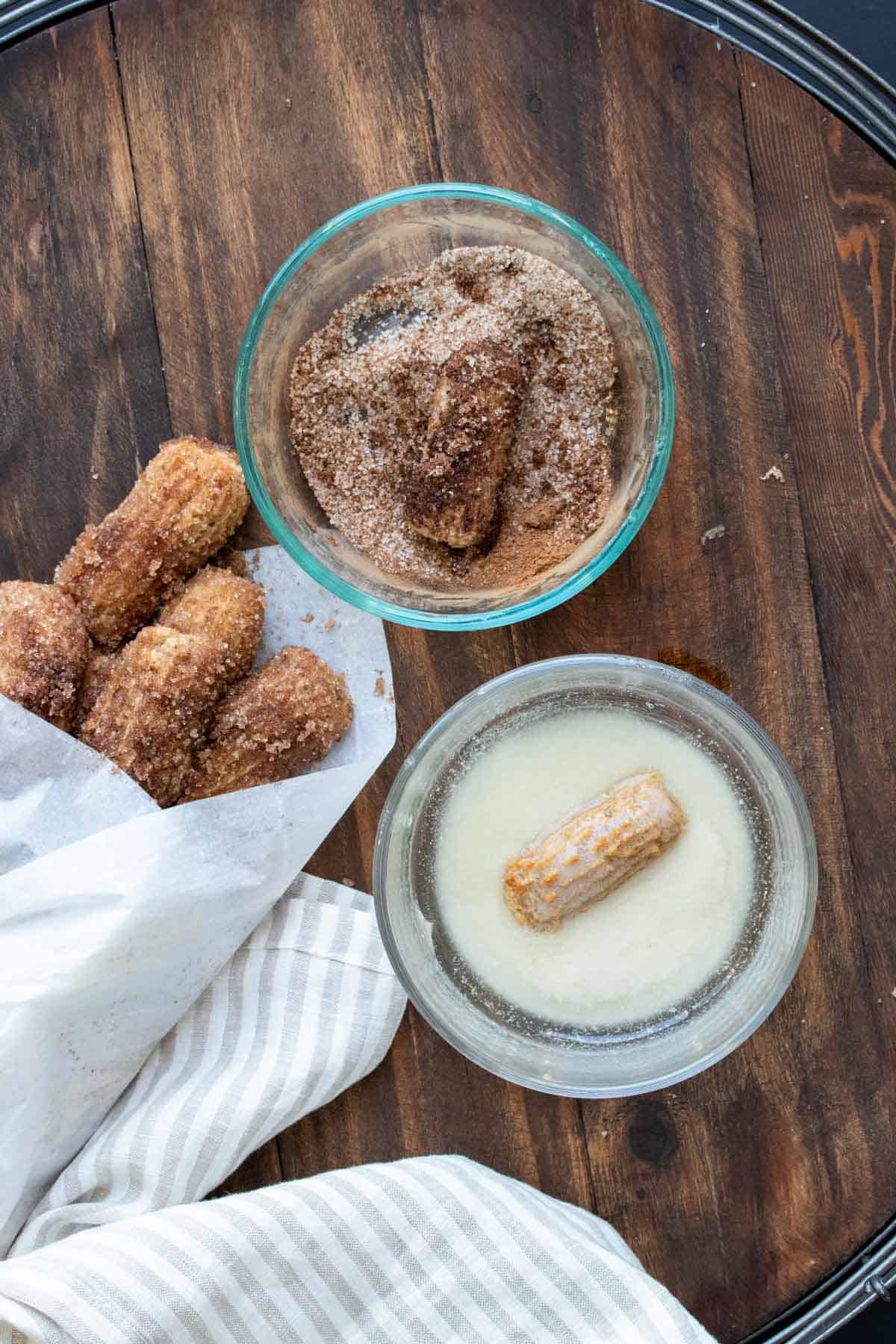 Churro pieces being dipped in a bowl of butter and then rolled in a bowl of cinnamon and sugar