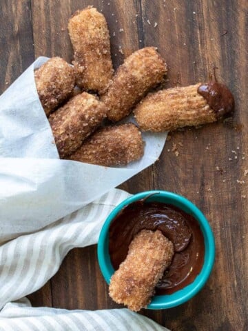 A cone of parchment paper filled with churro bites next to a bowl of one in chocolate.