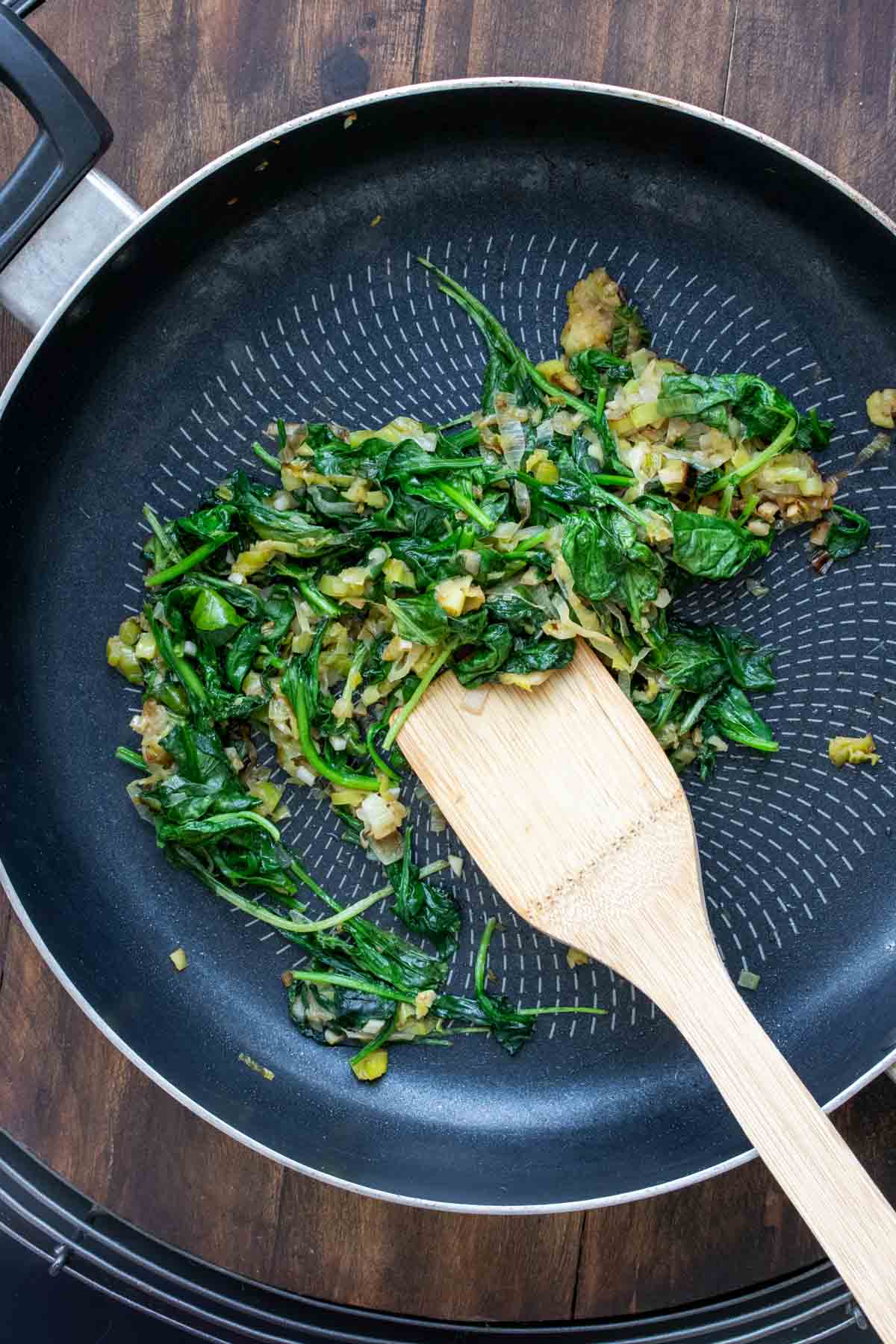 Wooden spoon mixing greens in a pan as they cook.