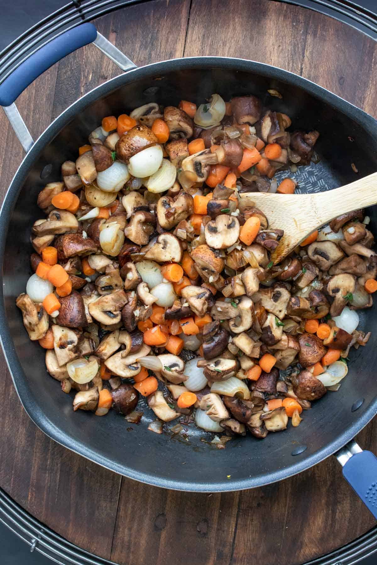 Carrots, mushrooms and onions being mixed in a pan by a wooden spoon