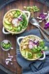 Two white soup bowls with pozole verde soup loaded with veggies toppings on a wooden surface.