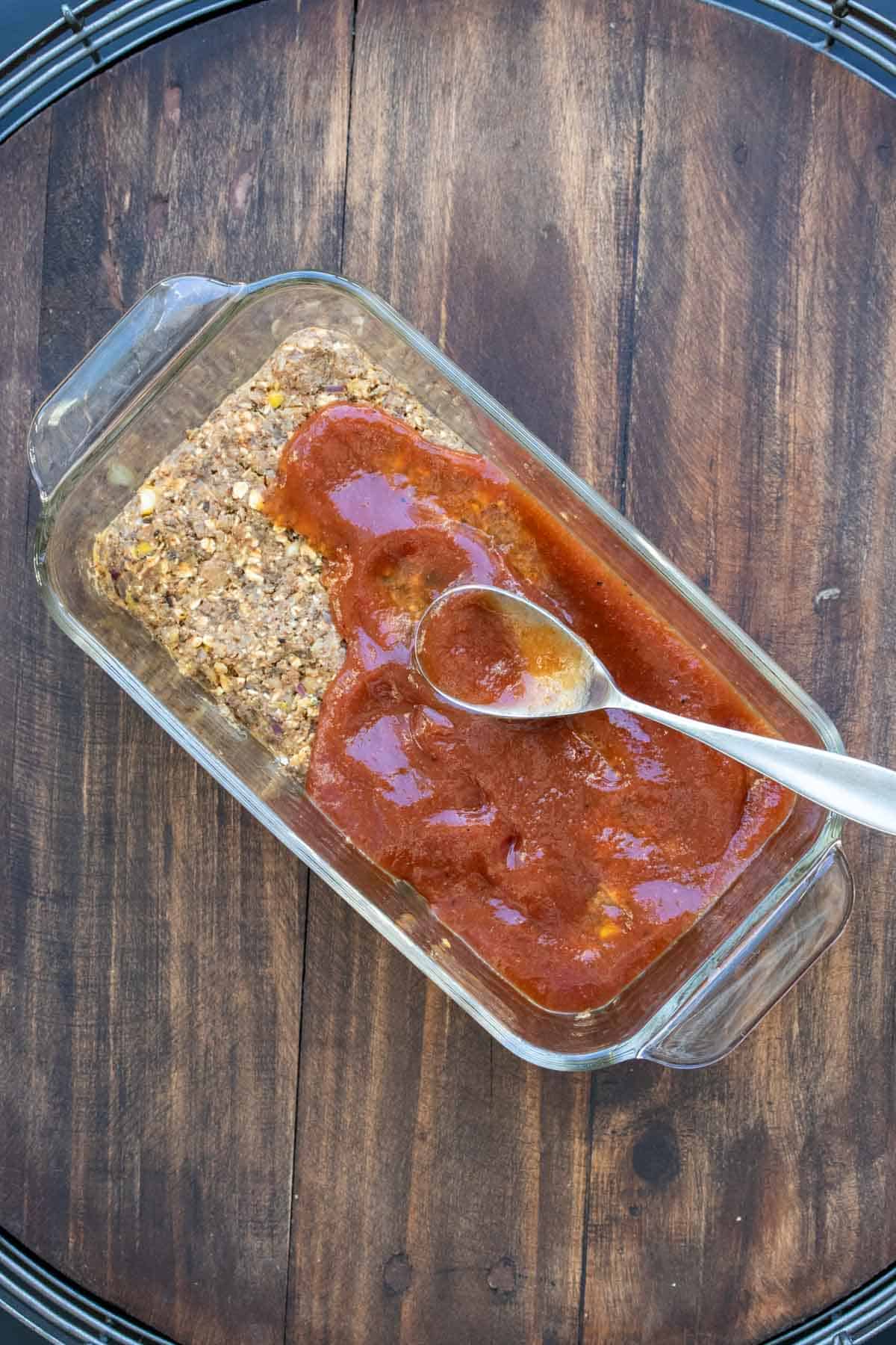 A spoon spreading barbecue sauce on lentil meatloaf mix in a glass pan