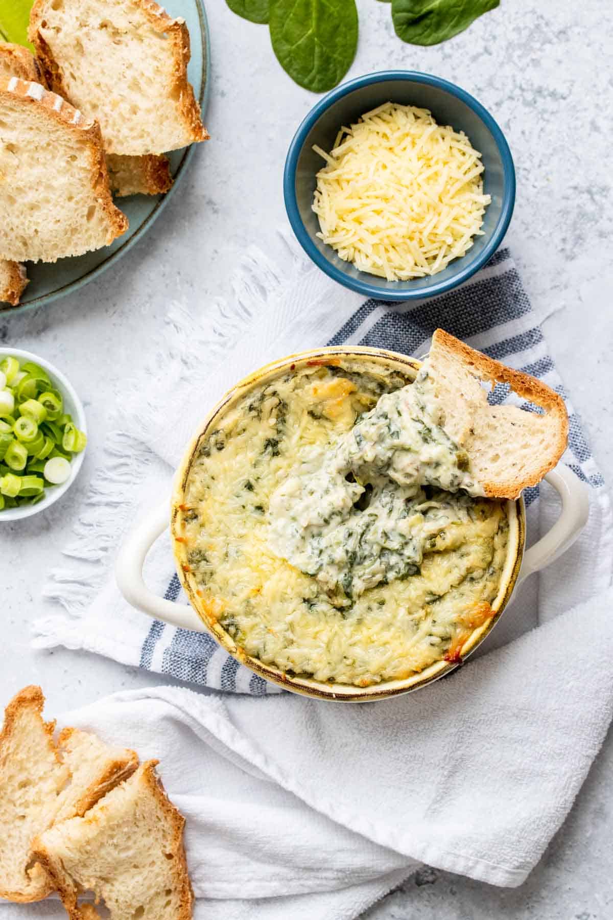 Top view of a bowl of baked spinach dip with a dipped piece of bread sitting on the edge next to a bowl of Parmesan