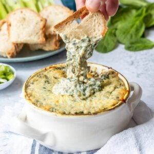 Spinach artichoke dip in a white ceramic bowl with toasted bread dipping into it.