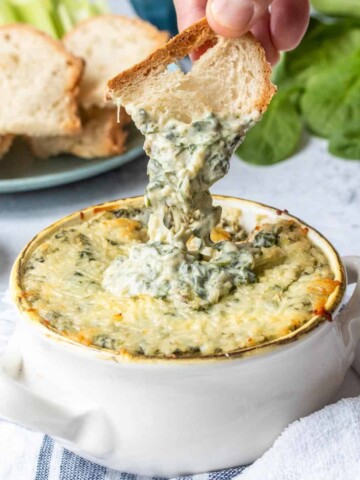 Spinach artichoke dip in a white ceramic bowl with toasted bread dipping into it.