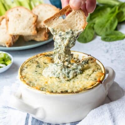 Spinach artichoke dip in a white ceramic bowl with toasted bread dipping into it