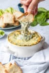 A hand dipping a piece of toasted bread into a baked spinach dip in a white casserole bowl.