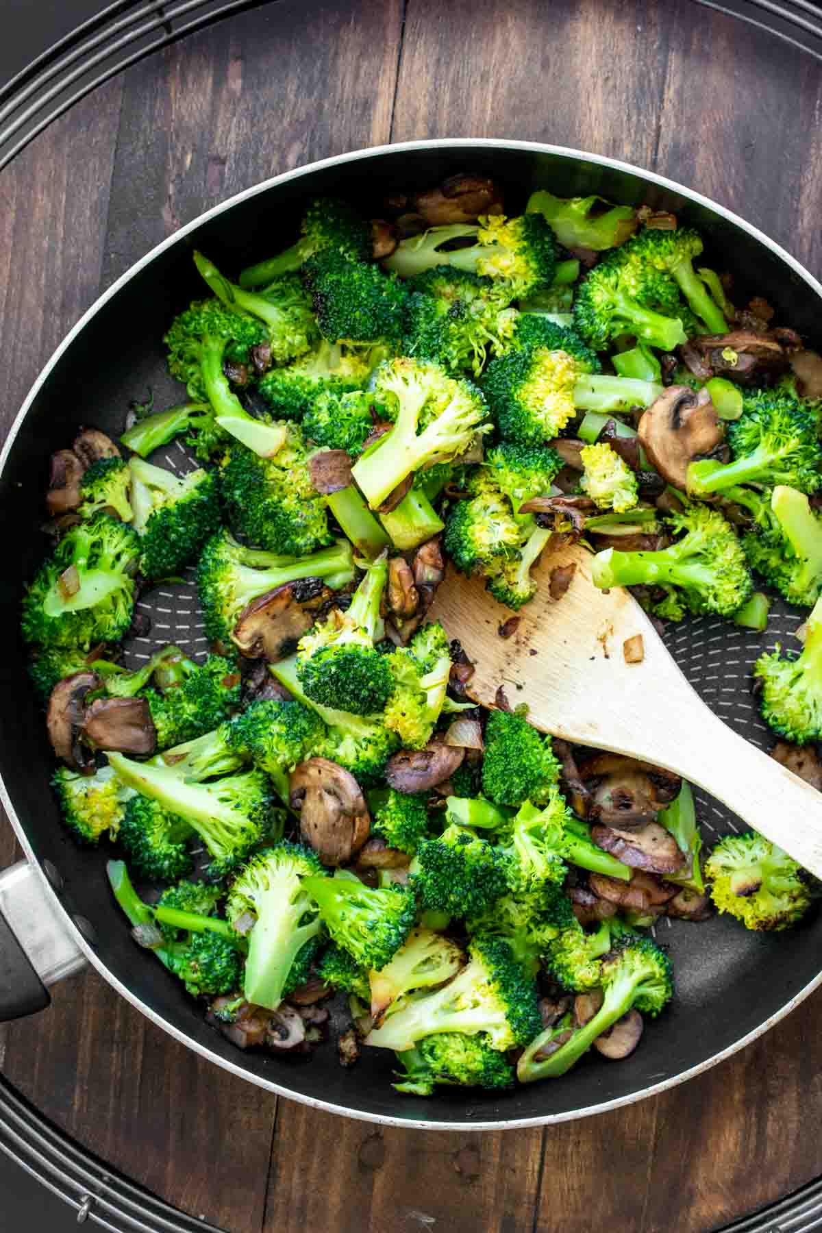 Broccoli and mushrooms being sautéed in a pan with a wooden spoon.