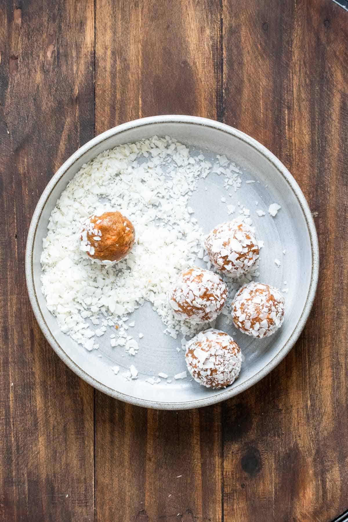 Carrot energy balls being rolled in shredded coconut on a grey plate