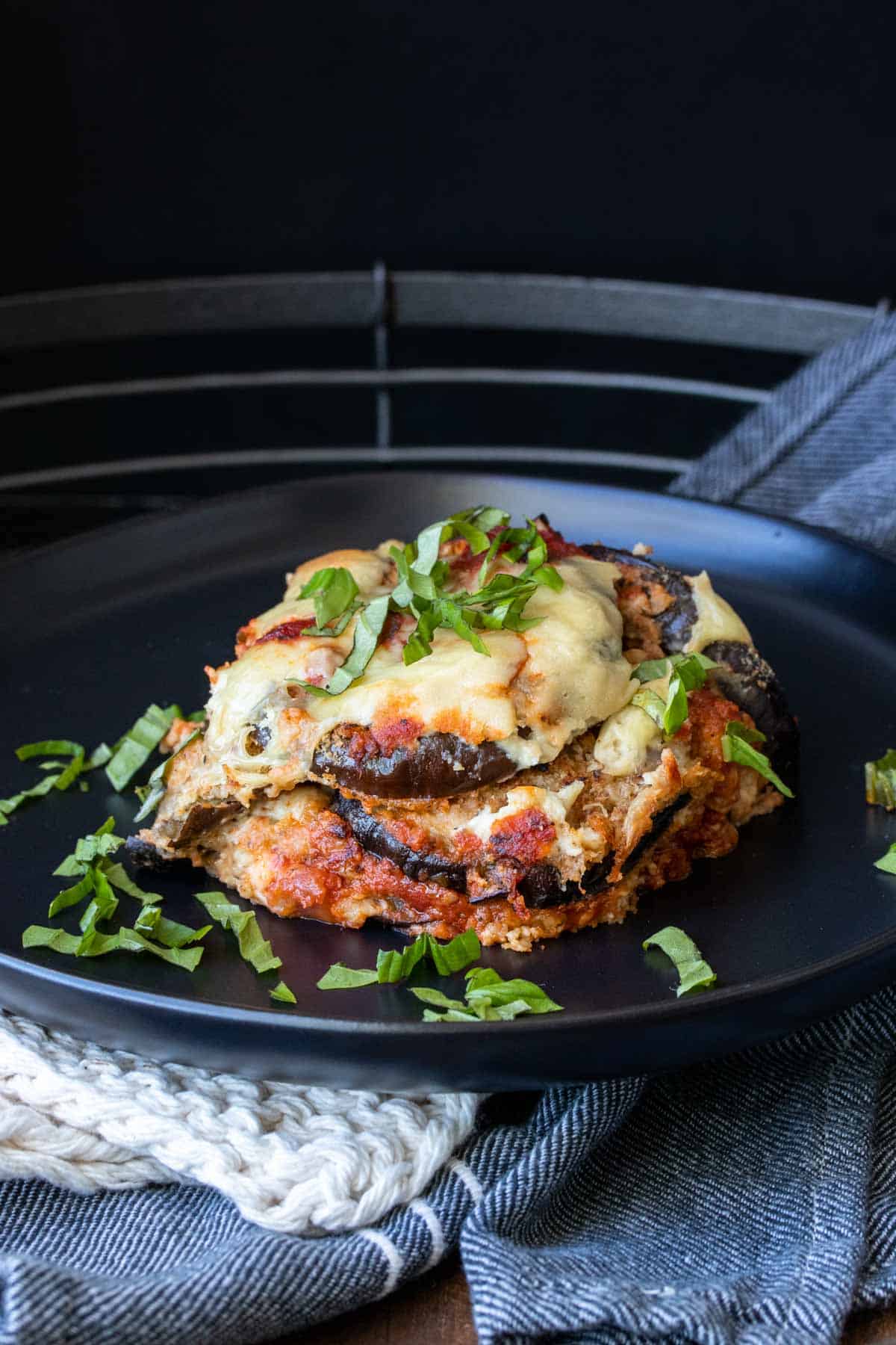 Black plate with a piece of eggplant parmesan topped with sliced basil