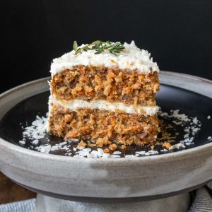A frosted piece of carrot cake on a black plate with shredded coconut on it.
