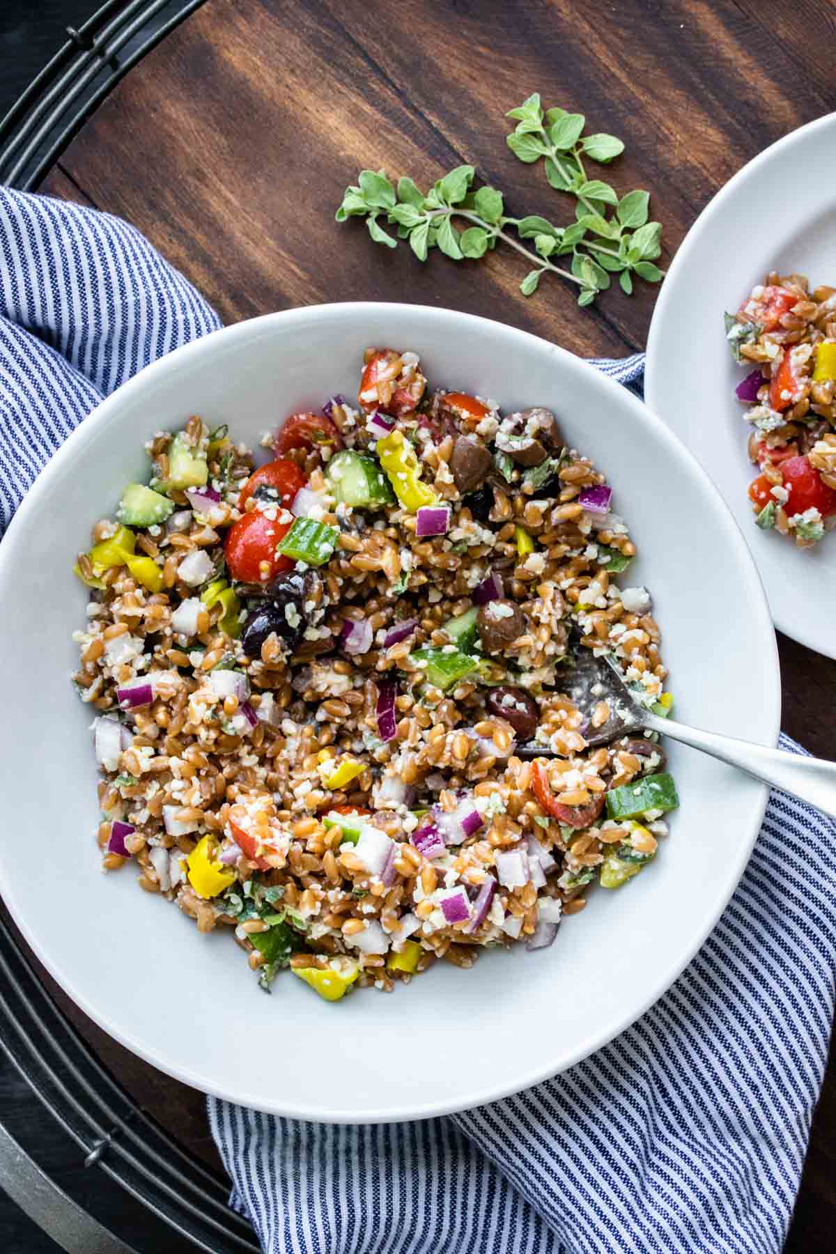 Spoon in white bowl filled with Greek farro salad on a blue and white striped cloth.