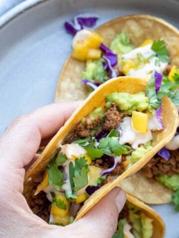 A hand holding a taco with lentil meaty crumbles, avocado and mango salsa