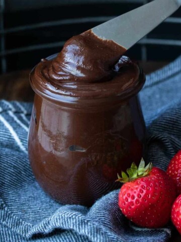 A knife getting Nutella out of a jar sitting on a blue towel next to strawberries