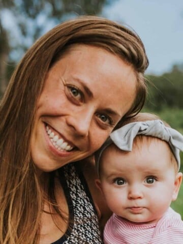 A mom with long brown hair smiling and holding her baby with a bow