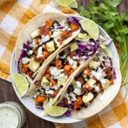 Three sweet potato and black bean tacos on a white plate sitting on a yellow checked napkin