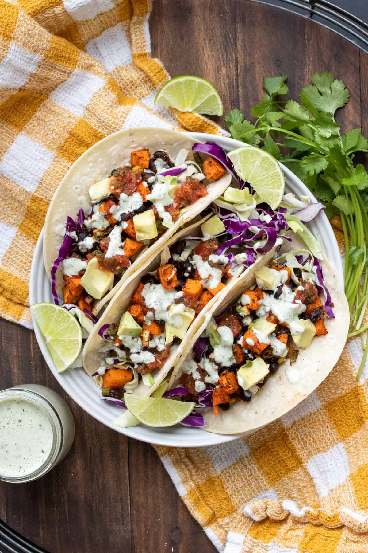 Three sweet potato and black bean tacos on a white plate sitting on a yellow checked napkin