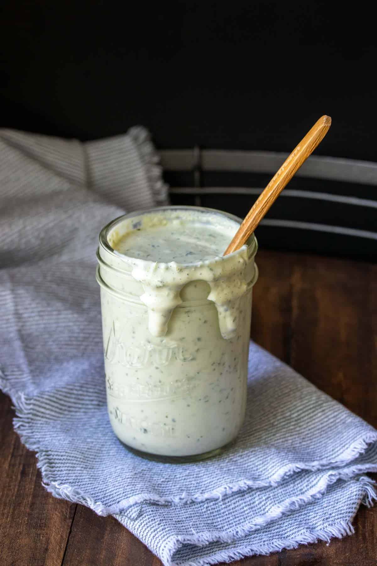 A glass jar with a creamy herb sauce sitting on a napkin on a wooden surface
