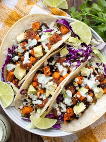 A white plate filled with three sweet potato and black bean tacos covered with white sauce and cabbage