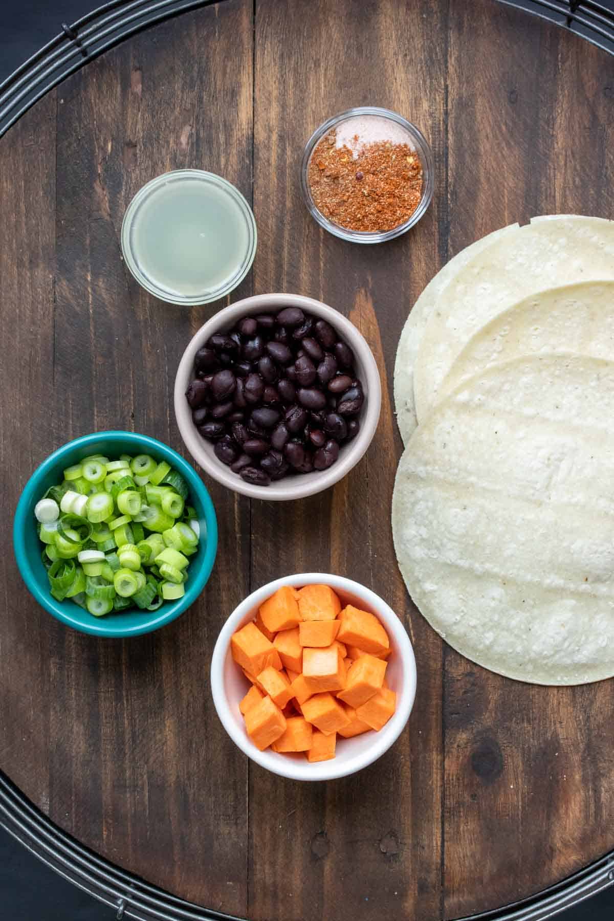 Bowls with chopped sweet potatoes, black beans, green onions, lime juice and spices next to a pile of corn tortillas