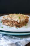 A piece of moussaka sprinkled with parsley on a white plate that is sitting on a blue plate.