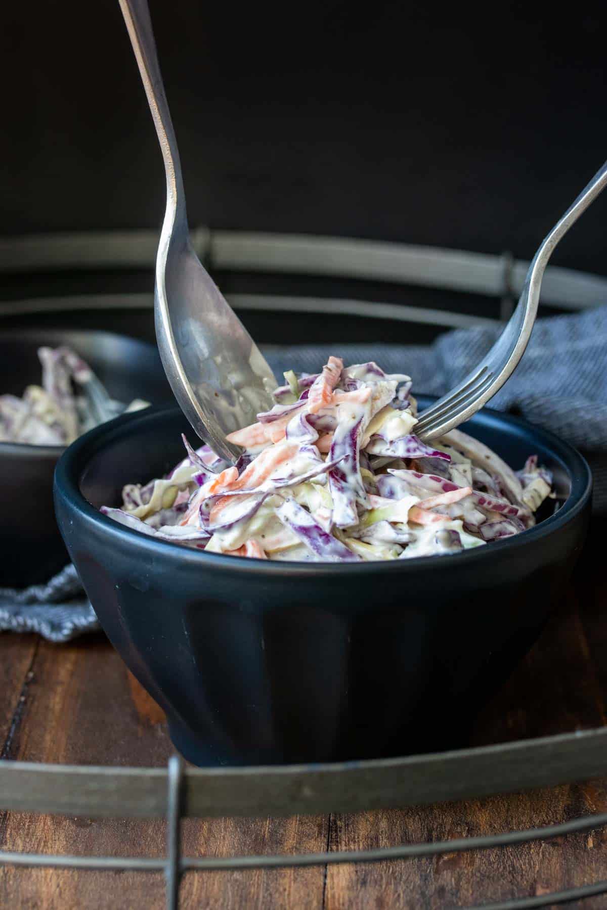 Two forks getting creamy coleslaw out of a black bowl.