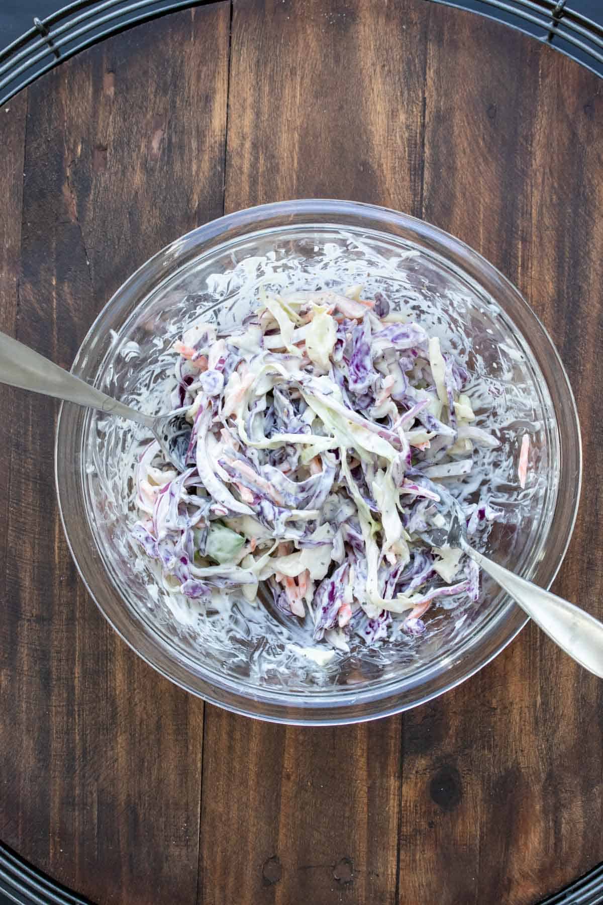 Glass bowl with forks mixing coleslaw inside.