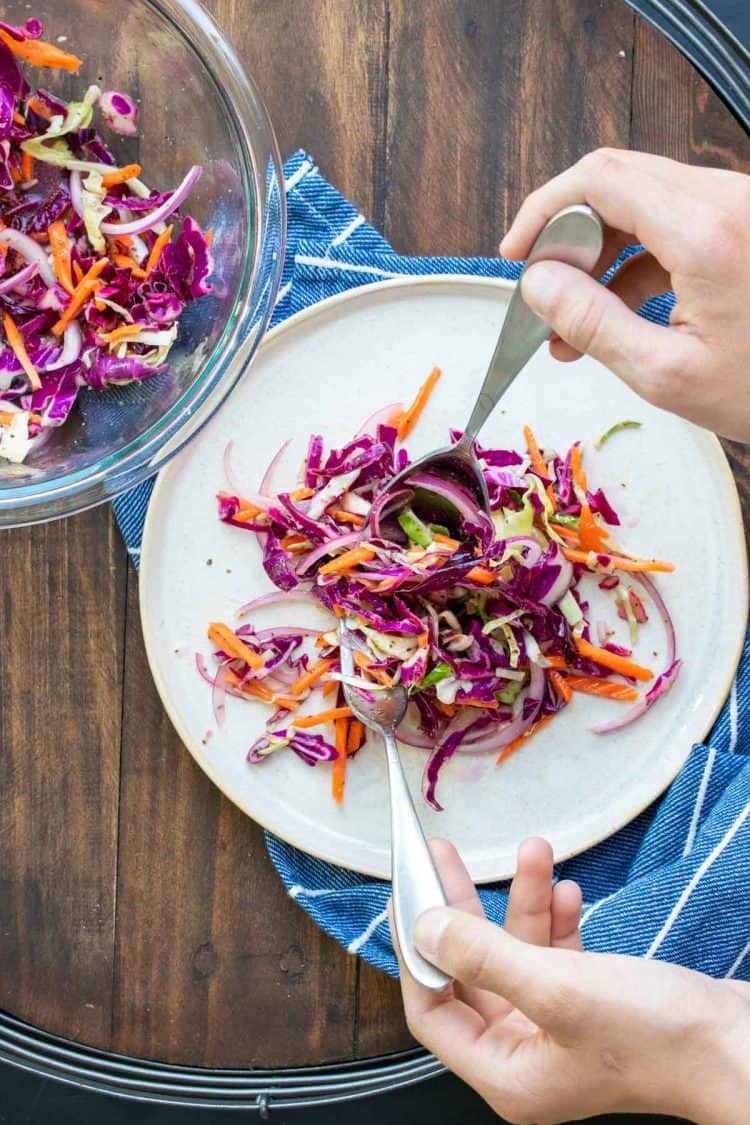 Hand holding two forks dropping cabbage slaw on a white plate.