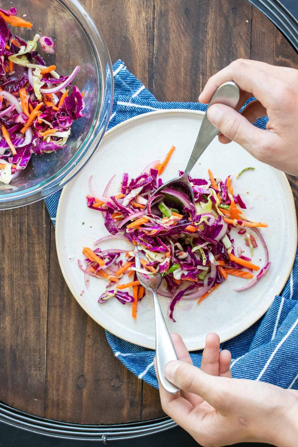 Hand holding two forks dropping cabbage slaw on a white plate