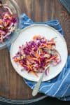 Vinegar cabbage slaw on a white plate sitting on a blue towel with white stripes next to a glass bowl of more slaw.