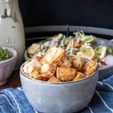 Side view of a grey bowl filled with baked croutons on a tray with salad and dressing