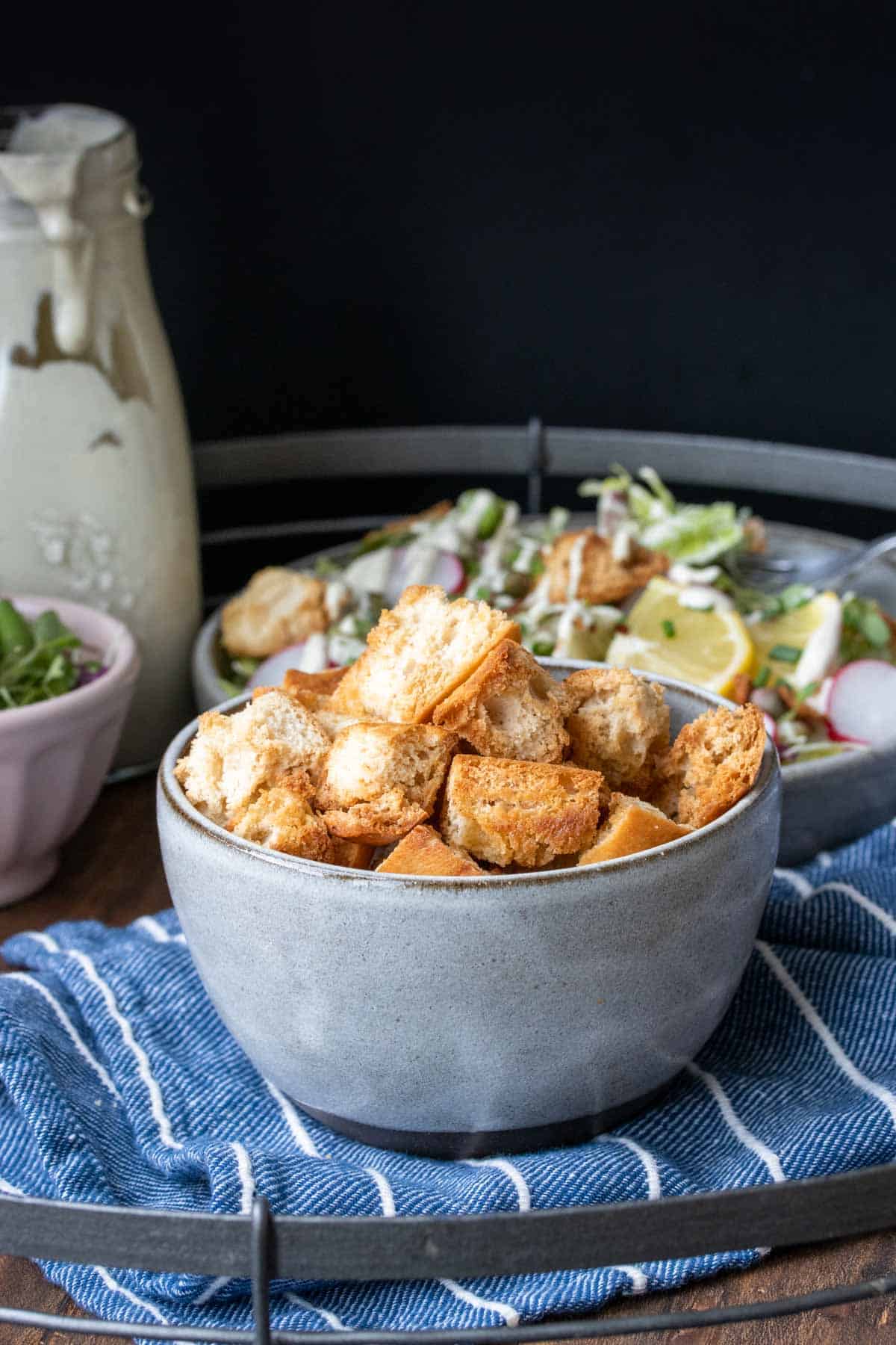 Side view of a grey bowl filled with baked croutons on a tray with salad and dressing